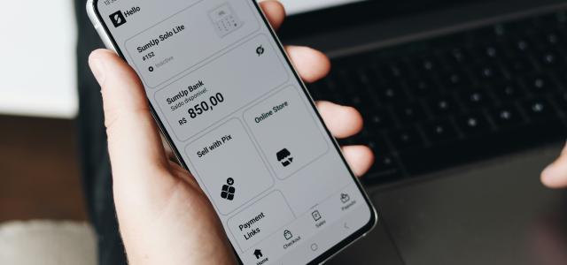 A person holding a cell phone in front of a laptop by SumUp courtesy of Unsplash.